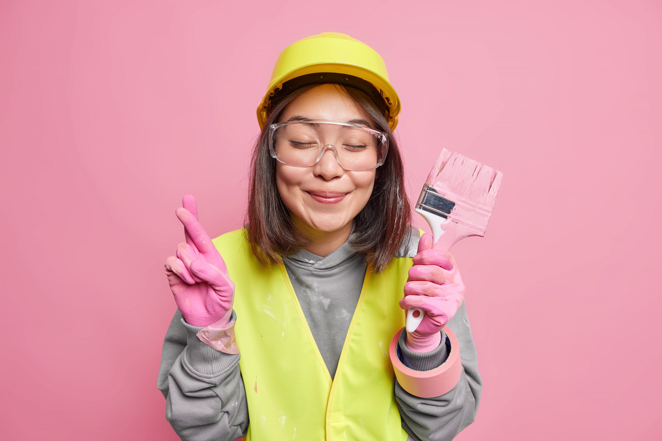 Pleased Asian woman crosses fingers holds paint brush renovates house makes wish believes in good luck wears safety glasses helmet gloves isolated over pink background works at construction company